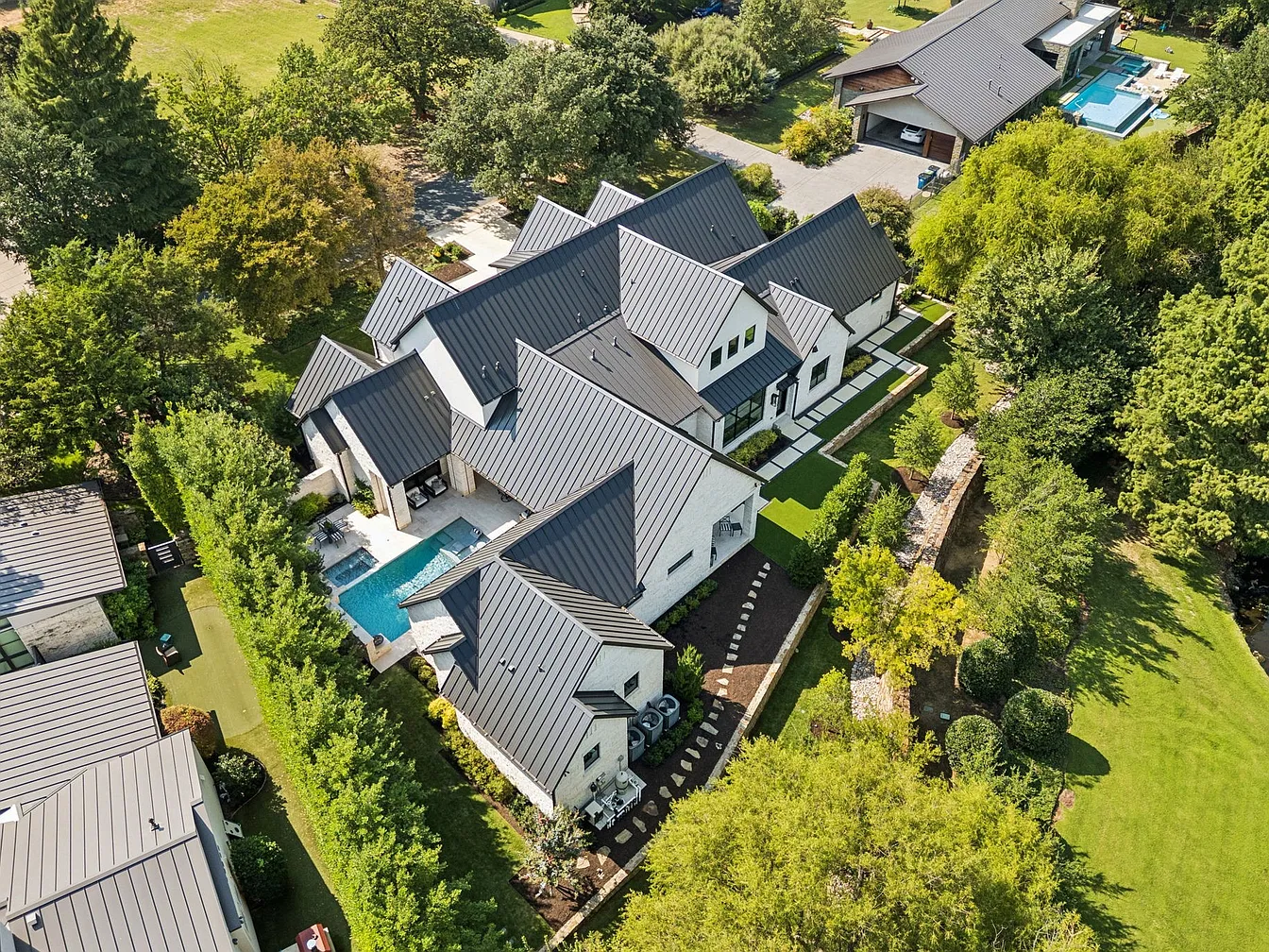 Aerial drone view of a modern contemporary estate in Westlake, TX 76262 with a new standing seam metal roof installation by RoofDog — white stucco and stone exterior, pool, and lush landscaping